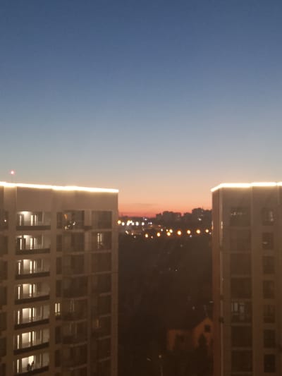 The image depicts an evening cityscape in Zhilino-1, Russia. Two tall apartment buildings, illuminated by warm, white strip lighting along their upper edges, frame the view. The building on the left has a prominent red light near its rooftop. In the middle ground, a silhouette of a town is visible with scattered yellow lights, suggesting a residential area or a street. The sky transitions from a clear blue at the top to a gradient of orange and pink near the horizon, indicating dusk or dawn. The overall impression is a residential urban environment during a transitional period of daylight.