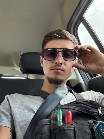 A man is pictured inside a vehicle, likely a car, wearing sunglasses and a grey t-shirt. He is touching his sunglasses with his right hand. A black bag with two items clipped to it, a green lip balm and a red marker, is visible in the foreground. The vehicle appears to be in a rural or suburban setting, not explicitly in the city of Biaga, Bulgaria. The lighting suggests it is daytime, possibly overcast.