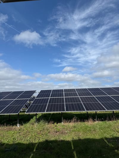 The image depicts an outdoor solar photovoltaic (PV) array situated in Southampton, United Kingdom. Multiple parallel rows of dark blue-black solar panels, each composed of a grid of individual cells, are visible across the midground. These panels are elevated by dark metal support structures, casting distinct shadows onto the ground beneath them. The ground cover consists of green grass and scattered weeds. The sky, which occupies the upper half of the frame, is bright blue with a mix of white clouds, including denser cumulus formations along the horizon and wispy cirrus clouds at higher altitudes. A single white contrail is discernible in the upper left quadrant of the sky. No individuals, animals, or other man-made structures are observed within the frame.