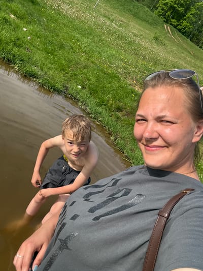 A woman and a boy are standing in a body of water with grassy shores in Novosinkovo, Russia. The woman is smiling and looking at the camera while taking a selfie. She is wearing sunglasses on her head, a gray T-shirt, and has a brown purse strap over her shoulder. The boy is wearing black shorts and a necklace with a cross. The water appears to be murky. The grassy shoreline is visible on the left of the frame.