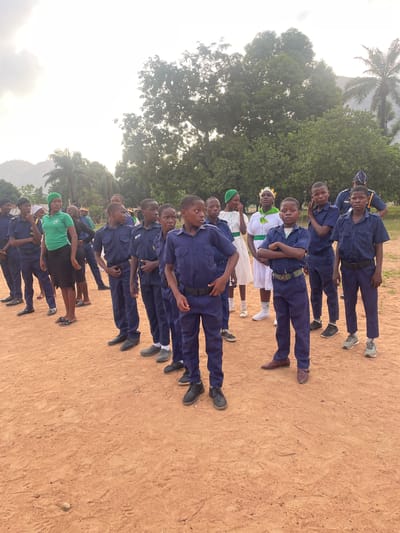 A group of approximately 15 people, mostly young males, are standing in rows on a dirt ground in Andaha, Nigeria. The majority of the group is wearing matching dark blue uniforms with belts and dark shoes. Several individuals in the middle rows wear white uniforms with green sashes and green head coverings. They all appear to be standing at attention. The environment includes green trees, a palm tree, and possibly a mountain in the background under a slightly overcast sky.