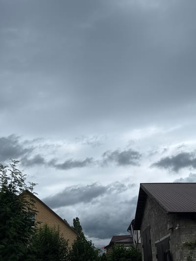 The image depicts a cloudy sky over a residential area. In the foreground, on the right, is the corner of a dark-roofed building constructed from gray cinder blocks. To the left of this building, a portion of another building with a lighter-colored brick facade and a dark roof is visible. Dense green foliage obscures the lower parts of both buildings. The sky is filled with overcast, gray clouds, with lighter patches suggesting diffused sunlight. The scene appears to be a residential street or backyard. No people or specific actions are identifiable in the image.