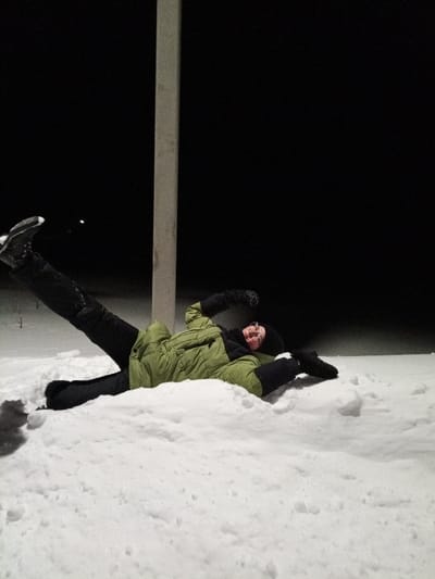 An individual lies supine in deep snow at night in Старый Шагирт, Russia. The person, an adult, is dressed in a light green winter jacket, black pants, black boots (with the right boot showing white sole details), a black knitted hat, a black scarf, and black mittens. Their right leg is extended upwards and angled to the left. The right arm is bent at the elbow with a clenched fist raised, while the left arm is bent with the hand resting on the snow. The individual wears glasses and looks towards the camera with a smile. A tall, light-colored, rectangular pole stands vertically behind the person, slightly to the left of the frame's center. The immediate ground is covered entirely by undisturbed white snow, with a dark background featuring a single faint light source visible in the far upper left. The foreground is well-lit, indicating a localized light source.