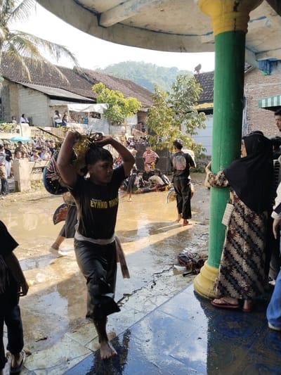 The scene depicts an outdoor event in Jenawi, Indonesia, evidenced by the traditional architecture of the buildings and the attire of the individuals present. A man in the foreground is carrying a basket-like object on his head, and he's wearing a dark t-shirt with text, dark pants, and a light-colored sash around his waist. He is barefoot and walking in what appears to be a puddle of water.

Behind him, there's a person wearing a similar dark t-shirt and pants, also barefoot. To the right, a woman in a black hijab and a patterned dress is leaning against a green and yellow pillar, possibly part of a covered structure.

In the background, there is a crowd of people observing the event. The ground is wet and reflective, suggesting recent rain. The environment includes traditional houses with tiled roofs and a backdrop of hills or mountains. The scene seems to capture a cultural event or ceremony. A person is sitting on the roof of one of the buildings.