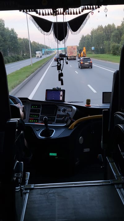 The image is taken from the interior of a bus or truck, looking forward through the windshield. The view shows a highway with multiple lanes of traffic, flanked by trees. Several vehicles are visible on the road, including a white truck, a yellow excavator, a light-colored car, and a dark-colored car.

The interior of the vehicle features a dashboard with various controls, a gear shifter, and a GPS device mounted on the windshield. Decorative tassels and trinkets are hanging from the top of the windshield.

The image appears to have been taken on a highway in or near Gruzinskii, Russia, based on the road signs and the style of vehicles. The lighting suggests it is either early morning or late afternoon. There is no clear indication of people in the immediate foreground, other than a hand on the steering wheel. The environment is that of a transportation route.