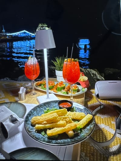 This image depicts an outdoor nighttime dining scene in Kumanovo, North Macedonia, situated next to a body of water. The foreground features a table covered with a yellow and white geometric patterned mat. On the table, from left to right, are: a dark speckled ceramic plate holding a folded white napkin and a silver fork; a small cylindrical metal container with white paper tissues; a stemmed glass containing a vibrant red cocktail garnished with a lime wedge and two dark straws; and a white, modern table lamp with a rectangular shade providing central illumination. To the right of the lamp, another stemmed glass holds an identical red cocktail, garnished with an orange slice and two dark straws, positioned in front of a plate with multiple portions of cucumber slices topped with a reddish substance and small fried elements. Further to the right on the table, a light gray purse with a metallic chain strap rests. In the immediate center foreground, a dark speckled plate presents several golden-brown, rectangular fried food items on a bed of green leaves, accompanied by a small black ramekin containing a red sauce. The background reveals a dark body of water reflecting distinct blue lights from a large, distant structure, possibly a bridge or building. A smaller, dimly illuminated object, resembling a boat, is visible on the water further to the right. A dark, rocky shoreline is visible between the table and the water. No individuals are present in the scene.