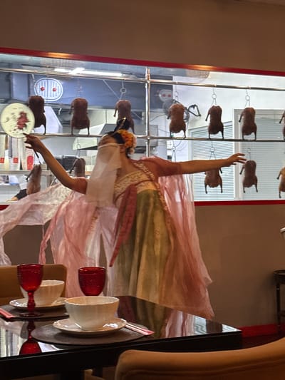 A person dressed in traditional East Asian attire, including a flowing pink veil and a layered dress in green and white, is captured in mid-performance or dance. The individual holds a circular fan in their left hand, with their arm extended. Behind them, a display features numerous roasted ducks hanging on hooks, illuminated by overhead lights. The background includes what appears to be a commercial kitchen setting with stainless steel surfaces and ventilation systems. In the foreground, a dark table is set with white bowls, saucers, red wine glasses, and chopsticks, suggesting a dining area within the establishment. The lighting is dim, creating a dramatic atmosphere, and the image seems to be taken indoors.
