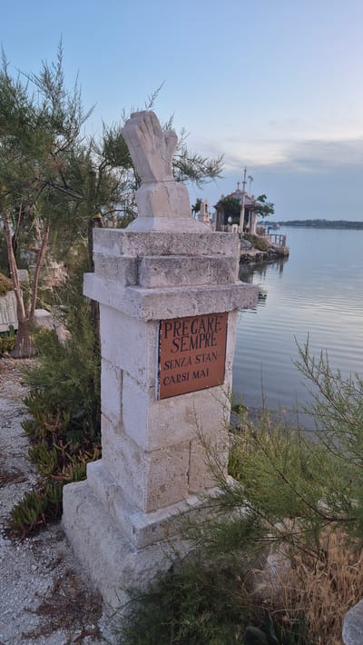 The image shows a stone monument with a plaque attached. The monument is weathered and appears to be outdoors, situated on a rocky outcrop overlooking a body of water. In the background, there are several small structures, some with crosses on top, suggesting a religious or memorial site. The water is calm, with gentle ripples reflecting the sky. Lush green vegetation, including trees and succulents, surrounds the monument.
The plaque on the monument bears an inscription in Italian. The text reads: "PREGARE SEMPRE SENZA STAN-CARSI MAI". This translates to "PRAY ALWAYS WITHOUT EVER GETTING TIRED". The monument itself is roughly carved stone, with a depiction of clasped hands or a similar gesture carved into its side. The overall impression is one of solemnity and peace.
The lighting suggests it is either early morning or late evening, with a soft, diffused light illuminating the scene. The sky is a pale blue with wispy clouds. The location appears to be coastal, given the presence of the sea and the hardy vegetation. The context provided by the prompt indicates the location is Taranto, Italy, which is a port city in southern Italy. The scene evokes a sense of contemplation and devotion, fitting for a place of prayer and remembrance.