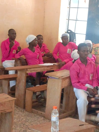The image displays a classroom scene in Mada, Nigeria, featuring approximately eight young individuals, presumed students, seated at wooden desks and benches. All visible individuals wear a uniform of bright pink long-sleeved shirts with emblems on the left chest pocket and white lower garments; some female students also wear white head coverings. A male student on the far left holds a small white object to his mouth, while another student in the mid-right holds a yellow pen. Various students exhibit neutral expressions, with some looking towards the camera and others in different directions. Papers and notebooks are present on several desks. A clear plastic water bottle with an orange cap stands on a wooden desk in the immediate foreground. The classroom has light-colored, possibly painted, walls with visible text markings, including "GULROY" and "GANG." A barred window, admitting natural light, is positioned on the right side of the frame, with a dark backpack visible beneath it. The floor features a mottled, light-toned pattern. One desk has "SAVE COCO UGA" visibly carved or written on it.