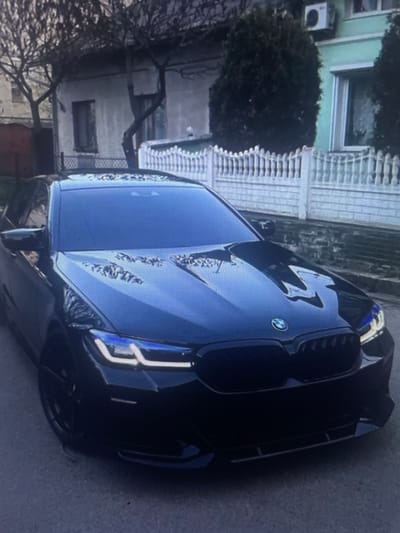 A black BMW sedan is parked on a street in front of a house. The car has tinted windows and distinctive blue and white headlights. The house behind the car has a light-colored facade and a white fence. Bare tree branches are visible in front of the house. The street appears to be paved with asphalt. The image is taken from a slightly elevated, frontal perspective. The location is the city of Kołobrzeg, Poland.