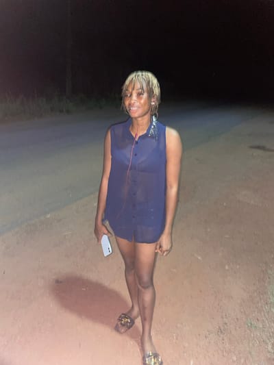 The image shows a woman standing on a dirt road at night. The location is possibly near Ogbe, Nigeria, based on available information. The woman is wearing a dark, translucent blouse and appears to be holding a phone. The surrounding environment consists of dark areas with potential vegetation or trees in the background, and a lighter-colored dirt surface under her feet. The overall lighting suggests the use of a camera flash or dim ambient light.