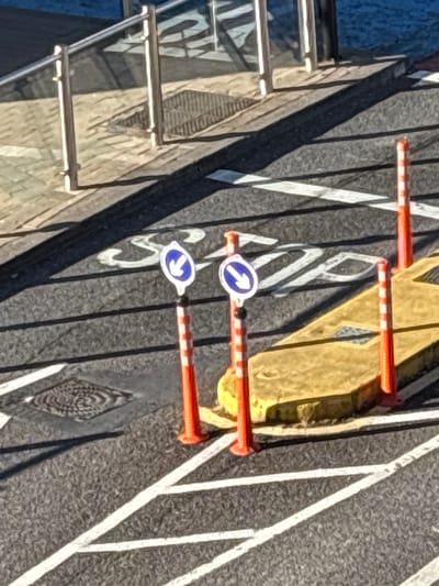 The image shows a road surface in Cork, Ireland, with traffic markings and street furniture. Two white arrows with blue circular backgrounds, indicating direction, are mounted on orange and white striped posts. These posts are positioned on a yellow concrete island which is adjacent to a pedestrian crossing. Further down the road, there are white line markings, some forming a pedestrian crossing, and a manhole cover. In the background, a metal railing and a shadow suggest the presence of a structure, possibly a building or a bridge. The scene is outdoors, on a paved road surface, under daylight conditions.