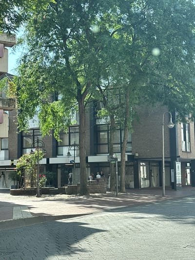 The image depicts a street view in Gütersloh, Germany, specifically the area near the event venue "Mondial". A multi-story building with a brick facade and large windows lines the street. Several mature trees with lush green foliage provide shade. In front of the building, a small fountain or planter area with rose bushes is visible. A streetlamp stands to the right of the fountain. Several people are seen in the vicinity: a group of individuals are seated on a low wall in front of the building, and others are walking or standing on the sidewalk. One person is visible inside a storefront. The ground is paved with cobblestones.
