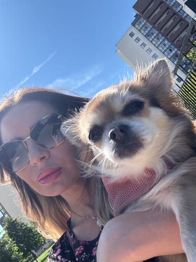 A selfie is taken outdoors on a sunny day with a clear blue sky. The photographer, a woman with blonde hair and sunglasses, holds a small, fluffy dog with tan and white fur. The dog is wearing a pink bandana. In the background, a modern, multi-story building with brown and white accents and many windows is visible. A black metal fence is also present. The environment suggests an urban setting, potentially a park or street in Saint Petersburg, Russia, given the architectural style of the building. The woman's lips are painted with a pinkish-red lipstick, and she is wearing a necklace.