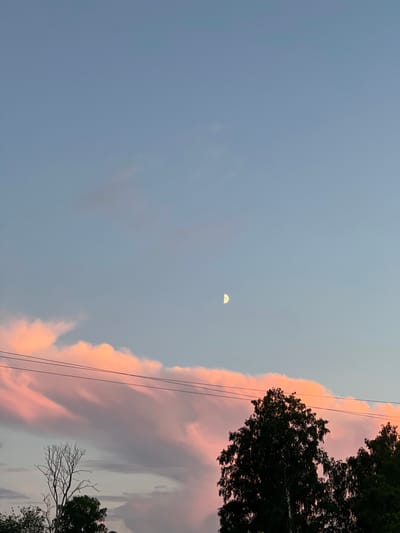 This is a photograph of a twilight sky over the city of Zaķumuiža, Latvia. The sky is a gradient of pale blue at the top, transitioning to lighter shades of pink and orange closer to the horizon, indicating the sun has recently set. A crescent moon is visible in the upper portion of the sky. Several wispy, pink clouds stretch across the middle of the frame. Three parallel dark power lines run horizontally across the image, intersecting the clouds. In the lower portion of the image, the dark silhouettes of several trees are visible against the brightly colored sky. One bare tree with intricate branches stands on the left, while a dense, leafy tree dominates the center and right. The overall scene depicts a tranquil evening landscape.