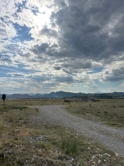 The image depicts a scenic view of a rural landscape with a dramatic sky. The dominant subjects are the sky filled with a mix of dark, heavy clouds and bright, sunlit ones, suggesting a dynamic weather pattern. Below the sky, a range of mountains forms the horizon, with a town or village nestled at their base, indicated by scattered buildings. The foreground consists of a rough, gravel path winding through dry, grassy terrain dotted with rocks and sparse vegetation. A single, dark-leafed tree stands in the middle ground, adding a vertical element to the otherwise horizontal composition.

The activity taking place is simply a view of the natural environment, with no people or specific events evident. The time of day appears to be late afternoon or early evening, judging by the quality of light and the shadows. The weather is mixed, with sunshine breaking through stormy clouds. The location cues, such as the mountainous terrain and the arid vegetation, combined with the known location context of Golubovci, Montenegro, suggest a Mediterranean or Balkan setting. The overall mood is one of natural beauty and tranquility, with a hint of dramatic atmosphere due to the weather. There is no visible text in the image.