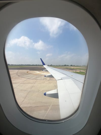 An airplane wing, predominantly white with blue tips, extends from the right side of the frame, occupying roughly half of the image. It is positioned over an airport tarmac, characterized by concrete slabs and yellow markings indicating taxiways. In the background, a line of trees and buildings marks the edge of the airport, with a clear blue sky dotted with white clouds overhead. The scene is viewed through an oval airplane window, its gray frame visible in the foreground. The location is identified as Vikram Nagar, India.