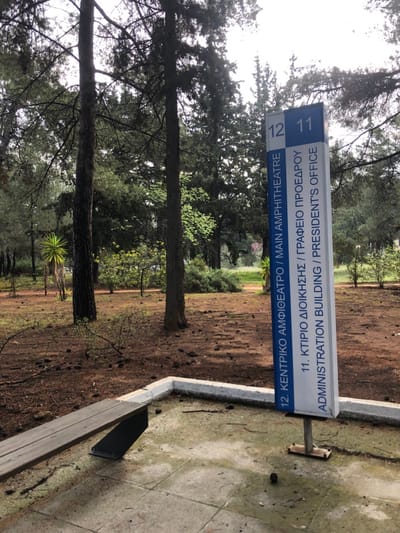 The image shows a signpost in the middle of a dirt path, standing in front of a bench. The sign is blue and white with black text and numbers. The signpost is located in a wooded area with trees and bushes in the background. The signpost indicates directions to two buildings: "12. KENTRIKΟ ΑΜΦΙΘΕΑΤΡΟ / MAIN AMPHITHEATRE" and "11. ΚΤΙΡΙΟ ΔΙΟΙΚΗΣΗΣ / ΓΡΑΦΕΙΟ ΠΡΟΕΔΡΟΥ ADMINISTRATION BUILDING / PRESIDENT'S OFFICE." The image is taken in the forest of Agia Paraskevi, Greece. The coordinates are latitude 37.999, longitude 23.82. The wooden bench is attached to a concrete base and faces the signpost. The signpost has a square, metal base. The ground is covered in dirt, rocks, and fallen pine needles. The sun is shining and there is a blue sky. The trees are green. There are no people in the image.