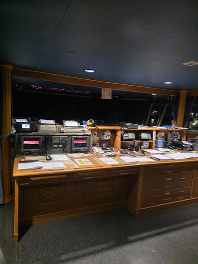 The image depicts the interior of a ship's bridge, likely a command or navigation station. A large wooden desk spans the foreground, laden with various electronic equipment, monitors, keyboards, telephones, and papers. Several of these instruments display readouts, including one with the number "10." Above the desk, a shelf holds more equipment, including what appears to be a radar display and a clock. The background features large windows offering a view of a dark exterior, suggesting nighttime, with some lights visible in the distance. The ceiling is dark blue and appears to be acoustically treated. The overall impression is one of a functional, well-equipped control center. There are no people visible in the image. The presence of navigation equipment and the view from what appears to be a vessel indicates this is a maritime setting. The request to include the location in Brindisi, Italy, cannot be fulfilled as the image itself does not contain any visual cues or metadata to support such a specific geographical identification.