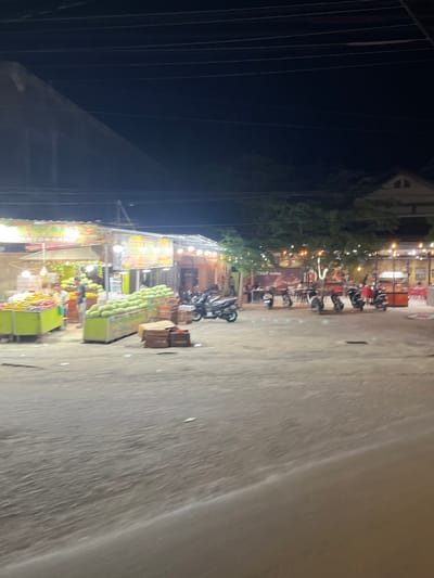 This is a nighttime, medium-shot, horizontal, slightly blurry photograph of an outdoor food stall area in Rawang Itiek, Indonesia. The scene depicts a row of food stalls with illuminated signs and hanging lights. In the foreground, the ground is a rough, greyish dirt or asphalt. To the left, there are stalls displaying piles of fruit, including what appear to be watermelons and corn, arranged in vibrant green and yellow bins. Two individuals, one wearing a blue shirt and the other in a darker shirt, are standing near the fruit displays. In the middle ground, several motorcycles are parked. Further back, more stalls are visible, some with tables and chairs, suggesting a dining area. One sign reads "TALLY Coffee." Trees and buildings are visible in the background, and power lines crisscross the dark sky above. The overall impression is of a lively evening marketplace.