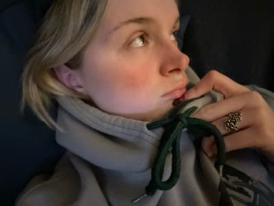 A fair-skinned person with blonde hair is shown from the chest up. The person is wearing a grey hooded sweatshirt. The hood's drawstring is dark green and tied. The person's right hand is visible, with a finger slightly touching their lip. A detailed, ornate silver ring adorns their finger. The person is looking off to the right of the frame. Their cheeks are flushed pink.