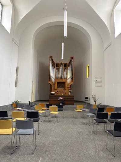 The image shows the interior of a church in Sesto San Giovanni, Italy. The main focal point is a large wooden organ with intricate carvings situated at the far end of the room, beneath a curved ceiling. Rows of chairs in various colors, including yellow, grey, and black, are arranged on the tiled floor facing the organ. Some chairs are empty, while others appear to be occupied by the back of unseen individuals. In front of the organ, there is a wooden altar with a book placed upon it, and floral arrangements. The walls are white, and the room is lit by overhead fluorescent lights and a small, yellow-tinted window on the right. The architecture features arched ceilings and large windows on the left. A sign with the number "8" is mounted on the right wall. The overall atmosphere is one of quiet reverence and a minimalist aesthetic.