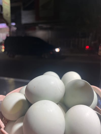 A close-up, low-angle shot shows a collection of white eggs in a pink woven basket. The eggs are mostly clean and have smooth shells, with one egg showing a faint hairline crack. The background is blurred and dark, suggesting an outdoor nighttime setting. Visible in the background are blurry lights that appear to be from streetlights and vehicle headlights, indicating a city environment. There is a dark silhouette of a vehicle in the mid-ground and possibly some buildings or structures further in the distance. The overall scene suggests a street-side stall or market in Kalijaten, Indonesia, during the evening or night.