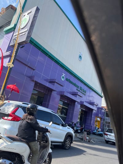 The image depicts a street scene in Jembrana, Indonesia, focusing on the facade of "Artasedana Market." The market building features a vibrant purple tiled lower section and a pale, ribbed upper section. A prominent sign with a stylized green fruit logo and the text "Artasedana Market fresh everyday" is visible on the purple facade. There are entrances with glass doors. To the left of the building, a tall yellow pole adorned with decorative elements and a small red umbrella is present.
In the foreground, a person wearing a black helmet and a black jacket is riding a white scooter. The scooter has "Scoopy" branding. A white SUV is parked to the right of the scooter. Several other scooters are parked further down the street in front of the market, and a silver car is visible on the right side of the road. The sky is blue with some clouds. The overall scene appears to be a daytime view of a commercial area.