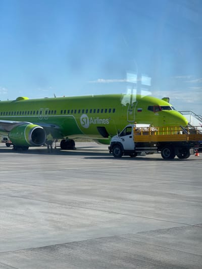 An S7 Airlines airplane is parked on the tarmac at Chulman Airport (UUDJ) in Chulman, Russia. The aircraft is predominantly green with the S7 Airlines logo on its fuselage. A small white utility truck with a yellow flatbed is parked next to the airplane. Two individuals, one wearing a yellow high-visibility vest, are standing near the plane's engines. The sky is clear blue with a few wispy clouds. The ground is paved with concrete.