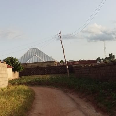 The image depicts a dirt road curving through a semi-urban setting in Mangu, Nigeria. The scene is bathed in the soft light of either early morning or late afternoon, indicated by the warm, low-angle sunlight. The sky is partly cloudy, with fluffy white clouds scattered across a pale blue expanse.
On the left side of the road, a light-colored concrete wall forms a boundary. Beyond it, a house with a distinctive corrugated metal roof, shaped like an inverted V, is visible. A lush green tree with dense foliage stands next to this house. Further down the road, a brick wall made of dark, rectangular bricks runs along the right side, separating the road from a cultivated area with rows of green plants.
A weathered wooden utility pole stands prominently in the middle ground, supporting two thin electrical wires that stretch across the sky. In the distance, the silhouette of a cell tower can be seen against the sky, along with other structures with red and corrugated metal roofs, suggesting a populated area. The overall impression is one of a rural or semi-rural community in Nigeria, likely during a calm period of the day, with no discernible activity or human presence visible in the frame. The dirt road suggests it might be a less-trafficked area.