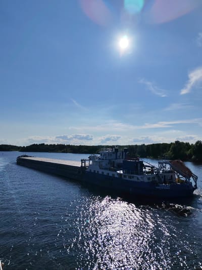 A barge is being pushed by a tugboat on a sunny day. The barge is a long, flat-topped vessel, and it is loaded with cargo. The tugboat is a smaller, more powerful boat that is used to tow or push large vessels. The barge and tugboat are on a wide river, with trees and vegetation on the banks. The sky is blue with a few white clouds. The sun is shining brightly, and its reflection can be seen on the water. The image appears to have been taken in Russia, as indicated by the Cyrillic text on the side of the tugboat, which reads "СЕСКАР" and "САНКТ-ПЕТЕРБУРГ". The time of day is likely midday, given the brightness of the sun and the clear sky. The weather appears to be warm and pleasant. The overall mood of the image is peaceful and serene, with the barge and tugboat moving smoothly through the water.