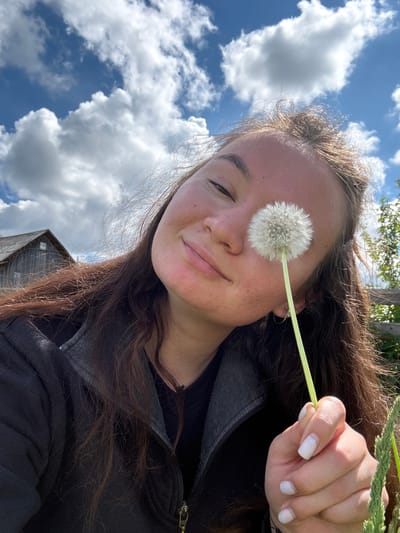 A young woman is holding a dandelion in front of her face. She has brown hair and is wearing a black jacket. The background is a blue sky with white clouds. There is a building in the lower part of the image. The picture was probably taken in Дойная, Russia.
