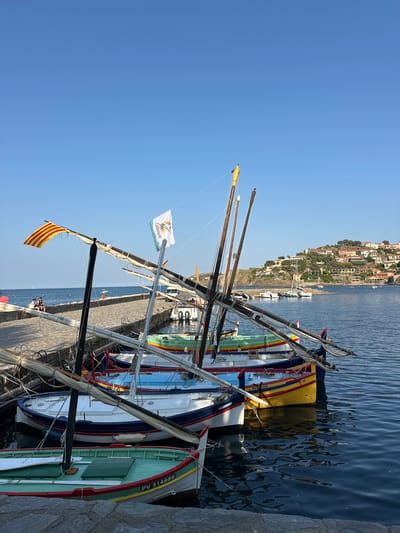 The image depicts a waterfront scene in Collioure, France. In the foreground, several colorful fishing boats are moored in the harbor. The boats are painted in vibrant hues of green, blue, yellow, and red. They are equipped with masts and rigging, suggesting they are traditional sailing vessels. Some boats have flags flying, including a Catalan flag and a flag with the word "Collioure" on it.

The boats are docked alongside a stone jetty. On the jetty, a few people are visible, some sitting and others standing. Beyond the boats and the jetty, the clear blue water of the harbor stretches out. In the background, the picturesque town of Collioure unfolds. Buildings with red-tiled roofs are clustered on a hillside overlooking the sea. A few other boats, including sailboats and a white motorboat, are anchored in the bay. The sky is a clear, cloudless blue, indicating a sunny day. The overall impression is one of a serene and charming Mediterranean coastal town.