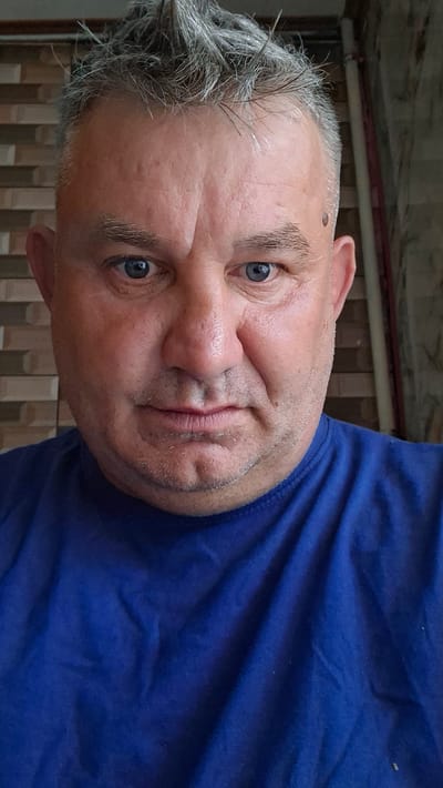 A man with short, spiky, grey hair and blue eyes is looking directly at the camera. He has fair skin with some redness, visible pores, and a mole on his right temple. He is wearing a blue t-shirt. The background consists of a brick wall and a vertical pipe. The image appears to be a selfie taken indoors. The setting is likely a residential area in Voinova, Moldova.
