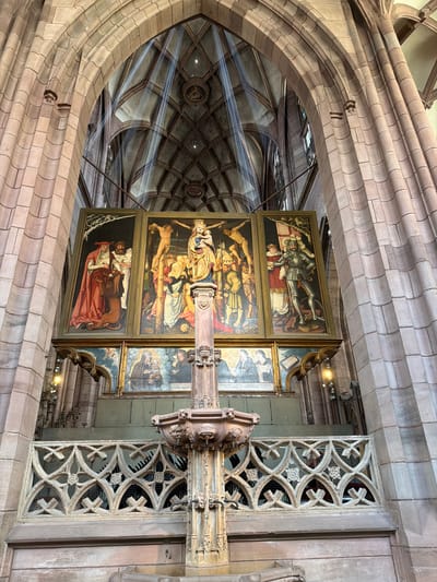 The image shows an interior view of a Gothic cathedral, likely Freiburg Minster, in Freiburg im Breisgau, Germany. The perspective is from a high vantage point, looking down and across the nave. The architecture features prominent stone arches, ribbed vaults, and intricate tracery.
Dominating the center of the frame are three framed paintings arranged vertically. The top painting depicts a figure in ornate armor standing over a seated figure in red robes. The middle painting shows a crucifixion scene, with multiple figures gathered around a central cross. The bottom painting features a scene with a figure in red robes lying down and a lion in close proximity. These paintings are part of a larger structure that includes carved elements and decorative grilles.
Light streams through the vaulted ceiling, creating dramatic diagonal beams across the space. The ceiling itself is a complex network of stone ribs forming diamond-shaped compartments, some of which contain roundels. The overall impression is one of grandeur and historical significance. The lighting suggests it might be daytime, with sunlight entering through unseen windows. The mood is solemn and reverent, fitting for a cathedral setting.