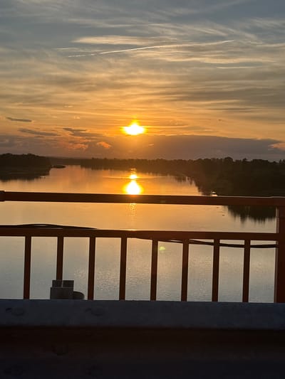 The image depicts a sunset over the Belaya River in Diurtiuli, Russia. The sun, a bright orange orb, is low in the sky, casting a warm glow across the water and illuminating the clouds. The river is wide and calm, reflecting the colors of the sunset. The banks of the river are lined with trees, and in the foreground, there is a bridge railing. The overall impression is one of tranquility and natural beauty.