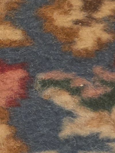 The image is a close-up, abstract view of a patterned textile, likely a carpet or rug. The dominant colors are shades of blue, beige, brown, and a touch of pink and green. The texture appears plush and somewhat fuzzy, suggesting woven fibers. The pattern is organic and floral, with swirling shapes and petal-like forms. No discernible objects, people, or environmental context related to Ladispoli, Italy, can be identified from this abstract close-up.