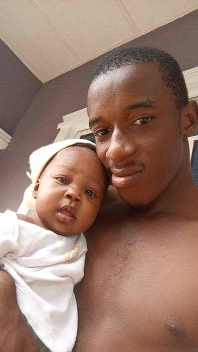 An image shows a shirtless, dark-skinned man holding a baby. The baby has dark skin and wears a white shirt and head covering. The man's head is slightly tilted towards the baby, and both subjects look forward. The background features a building's ceiling and wall. The man's complexion is clear, with short hair and minimal facial hair. The baby appears calm and alert.