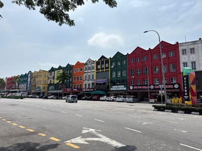 A row of colorful, multi-story buildings line a wide road. The buildings feature a uniform architectural style with gabled roofs and a mix of vibrant colors like red, green, yellow, and orange. Each building has storefronts at street level, marked by signage. Cars and a bus are parked along the curb. An "UPTOWN" sign stands at the end of the block. Trees overhang the buildings, partially obscuring the sky. The sky is mostly clear, with some clouds.