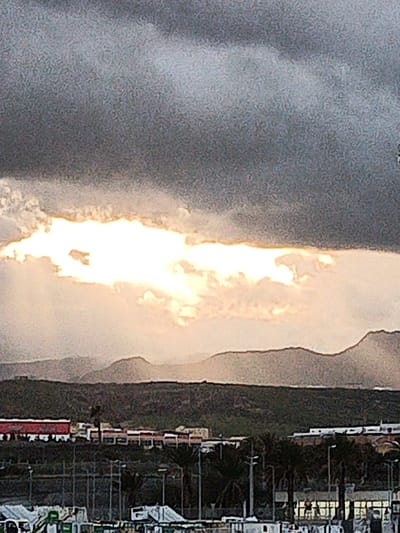 A cloudy sky dominates the scene, pierced by a bright, horizontal gap revealing the sun. Hazy mountains line the horizon. Below, a dark green expanse is populated with buildings, some appearing red-roofed. Palm trees and streetlights rise from the foreground, partially obscuring low, white structures. The light casts a warm glow on the landscape, contrasting sharply with the dark, overcast sky.