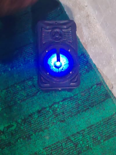 A dark, rectangular portable speaker is depicted in a dimly lit environment, resting on a green, textured mat or carpet. The speaker emits a bright blue light from its central circular element, illuminating the surrounding area and highlighting details on its grille. Small, light-colored rivets are visible around the glowing blue element, and geometric patterns are discernible on the speaker's dark-colored casing. To the upper right, a light-colored, possibly concrete or plastered, surface is visible, suggesting a wall or adjacent floor. No individuals, actions, or interactions are present in the static scene. This scene is located in the city of Yangikurgan, Uzbekistan.