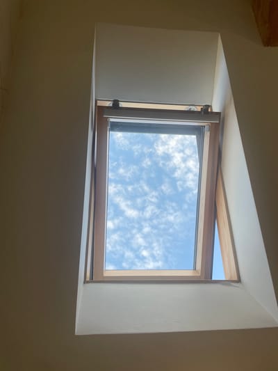 The image shows an open skylight with a wooden frame, looking up at a blue sky with scattered white clouds. The interior walls surrounding the skylight are painted a light color. The scene is indoors, likely in a residential or commercial building. No people or specific landmarks of Taganrog, Russia are visible.
