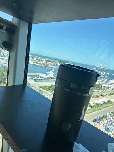 The image shows a view from a high vantage point in Gdynia, Poland, overlooking a marina and the Baltic Sea. In the foreground, a dark, cylindrical object, possibly a thermos or travel mug, is placed on a dark, flat surface. This object appears worn, with chipped paint. Through a large window, a bustling marina is visible, filled with numerous boats and yachts. In the background, a large sailing ship with multiple masts stands prominently in the water. Further out, the vast expanse of the blue sky meets the sea. The scene suggests a sunny day, with clear weather conducive to maritime activities. The architectural style of the buildings surrounding the marina suggests a modern urban setting.