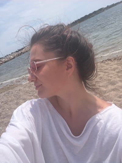 A woman with brown hair, tied back, is lying on a beach in Glifada, Greece. She is wearing a white top and pink sunglasses. The sea is to her left and sandy beach to her right. The image is rotated.