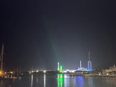 The image captures a nighttime view of a harbor in Agde, France, likely near the marina or a waterfront amusement area. The dark sky is punctuated by a single bright star or distant light in the upper left quadrant. The water in the foreground reflects the lights from the shore, creating shimmering streaks of color.
On the left side of the frame, several sailboats are docked, their masts reaching into the night. Their lights cast a warm glow on the water. In the middle ground, a collection of amusement park rides is illuminated by vibrant, colorful lights, predominantly green and blue. These rides appear to be tall structures, possibly roller coasters or observation towers, with bright vertical beams of light. A Ferris wheel or similar circular structure with blue lights is visible in the background.
To the right, a building with a white facade and a beige roof is partially visible, suggesting a residential or commercial area along the waterfront. The overall scene depicts a lively nighttime atmosphere with a blend of maritime activity and entertainment.