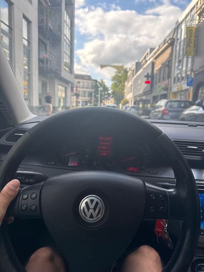 The image shows a first-person view from inside a car, looking out at a city street. The steering wheel of the car, a Volkswagen with its emblem visible, dominates the foreground. The dashboard displays various information on a red digital screen, including what appears to be speed (150 km/h), temperature (29.0°C), and potentially time (10:19). A hand is visible gripping the left side of the steering wheel.
Outside the car, the view is of a moderately busy street lined with buildings, likely shops, on both sides. The architecture suggests an urban European setting. Trees and a cloudy sky are visible above the buildings. A pedestrian is seen walking on the sidewalk in the middle distance. Cars are parked and in motion on the street. A yellow sign with "Panos" written on it is visible on one of the buildings. Based on information from other sources, this scene is located in Waregem, Belgium.