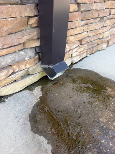A black downspout extends from an unseen source above and directs water onto a textured concrete surface. The downspout is positioned against a wall constructed of stacked, rough-hewn, tan and brown stones. A puddle of water has formed on the concrete, extending outwards from the downspout's outlet and appearing somewhat murky with traces of green algae or moss. The scene appears to be outdoors, likely on the exterior of a building in Cardwell, United States. There are no people or animals visible in the image.