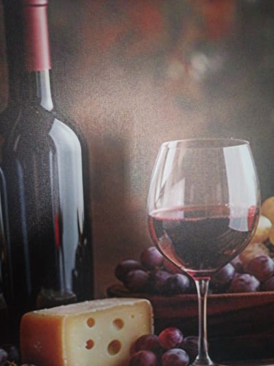 The image is a still life of wine, grapes, and cheese, set against a dark, textured background. A dark glass wine bottle with a red foil cap stands on the left. To its right, a stemmed wine glass is filled with red wine. In front of the glass, a wedge of Swiss cheese with visible holes sits on a surface, partially obscured by a cluster of dark purple grapes. More grapes are visible in the background, some in a wooden bowl. The lighting is soft and highlights the reflective surfaces of the glass and bottle. The overall mood is one of rustic elegance. The location in the city of Silifke, Turkey, is not discernible from the image.