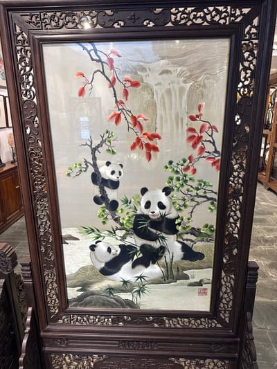 A framed silk embroidery artwork depicts three giant pandas interacting with bamboo and foliage, presented on a light-colored silk background. The pandas are rendered with detailed black and white fur patterns. One panda is climbing a tree branch laden with red leaves, while another sits and eats bamboo. A third panda is lying down in the foreground, partially obscured. The background features a waterfall or misty landscape. The artwork is housed in an ornate, dark wood frame with intricate carvings and a decorative base. A red seal stamp is visible in the lower right quadrant of the embroidery. The artwork is displayed indoors, with other furniture and artwork visible in the blurred background, suggesting an antique shop or gallery setting in Qingyang District, China.