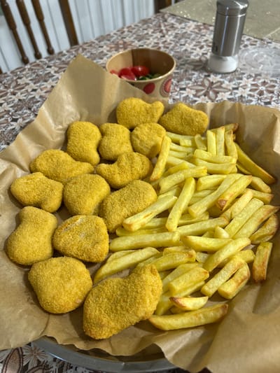 The image shows a close-up of a metal bowl filled with french fries and chicken nuggets, placed on a brown paper liner. The french fries are golden brown and appear to be freshly cooked, with some slightly darker, crispier pieces mixed in. The chicken nuggets are also golden brown and coated in breadcrumbs. They vary in shape and size, some being more rounded and others appearing more irregular. The bowl itself is a shallow, metallic dish.
To the left of the bowl, a small paper cup is visible, containing a few cherry tomatoes and some greenery, possibly a salad garnish. Behind the food and the cup, a patterned fabric or tablecloth is visible, with a floral design in shades of brown and white. A silver salt or pepper grinder with a white top is also in the background, positioned above the cup and fabric.
The lighting suggests it might be daytime, with no strong shadows indicating a specific time of day. The overall scene evokes a casual meal, perhaps served at home or in a casual dining setting. There are no people visible in the image. No text is clearly discernible on any of the items. The food is the primary focus, arranged invitingly within the bowl. The background elements provide context, hinting at a domestic or relaxed environment. The image is shot from a slightly elevated angle, looking down at the food.